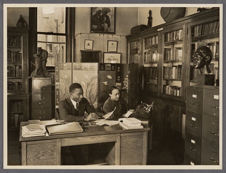 A sepia photograph, shown as a print on a pale gallery wall, of a library office. Curator Lawrence Reddick, in a suit and glasses, sits at a wooden desk covered with papers beside a woman reading. Behind them are glass-front bookcases, a painted folding screen, filing cabinets, and a sculpted head.