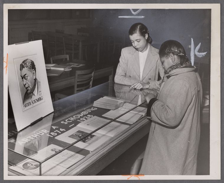A black-and-white photograph from 1952, shown as a print on a pale gallery wall. Two women lean over a glass display case lettered with Schomburg’s name and the years of his life. A drawn portrait of him stands at the left. One woman, in a light suit, points to a book, while the other, in a winter coat, looks closely.
