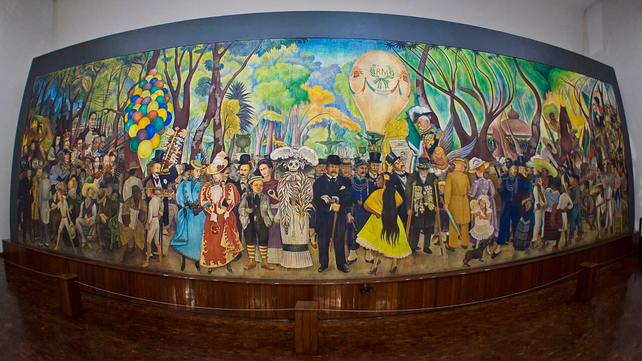 Diego Rivera's long, colorful mural of a crowd strolling through a tree-filled park, with bunches of balloons on the left and a hot air balloon rising above the center. At the very center stands La Catrina, a skeleton in a long white dress with a feathered boa and a hat crowned with plumes. Posada, in a black suit and bowler hat with a cane, stands at her left, and young Rivera, in a boater hat and striped stockings, holds her other hand, with Frida Kahlo just behind him. The wide-angle photo curves the mural slightly, with the museum's wooden base and rope barrier in front.