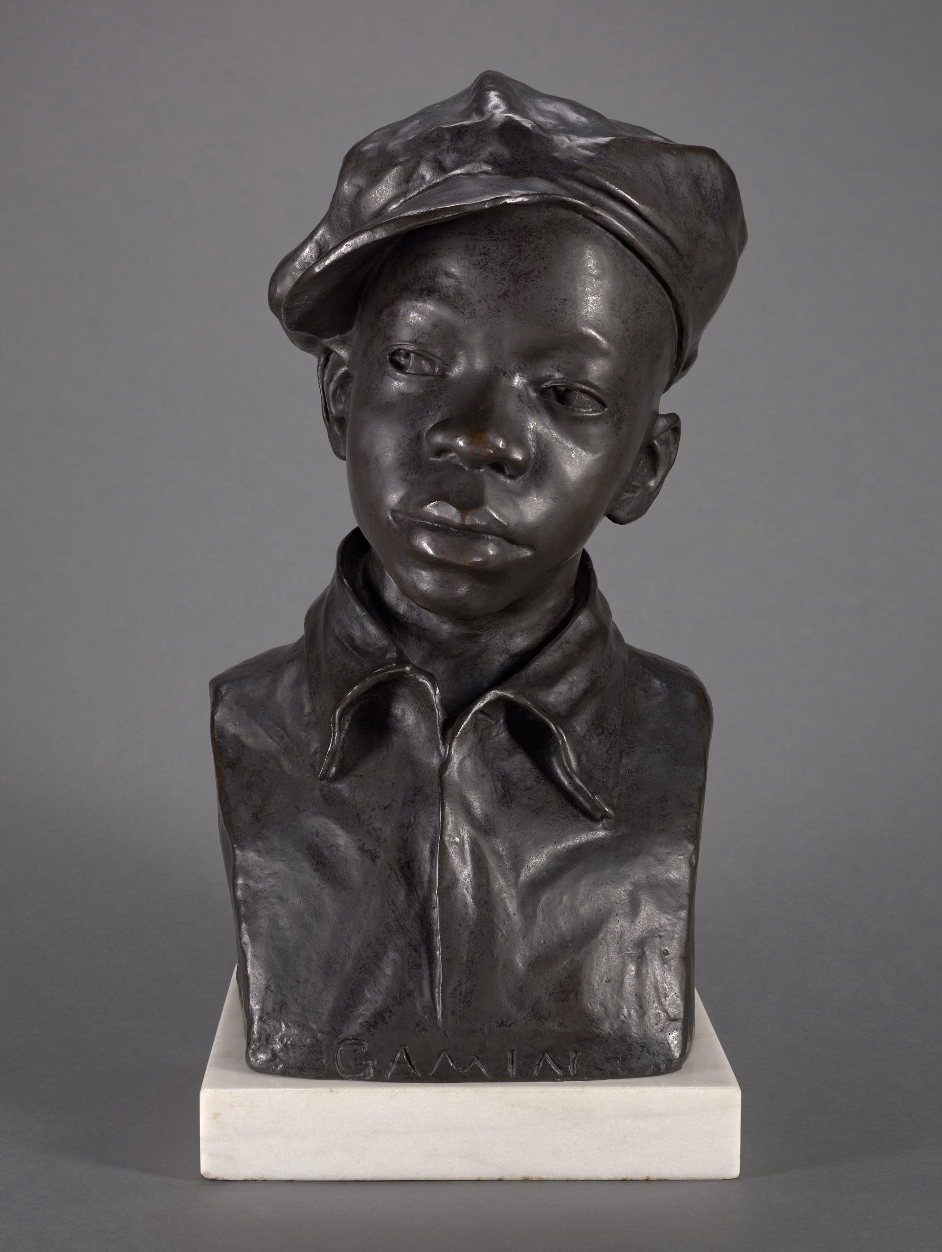 Sculpted bust of a Black boy in a soft cap tilted to one side, his gaze calm and steady, set on a white base.