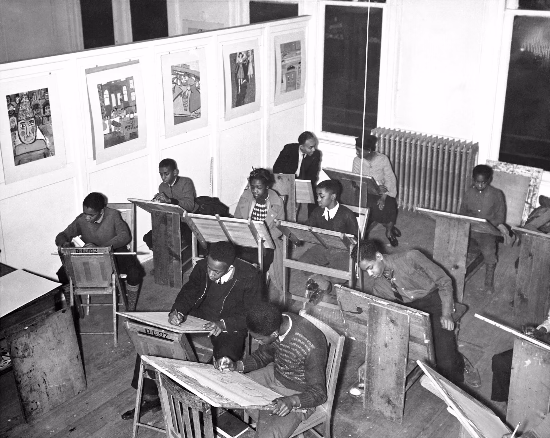 Black children and teens draw at slanted wooden boards in an art class, with two adults at the back and student paintings on the wall.