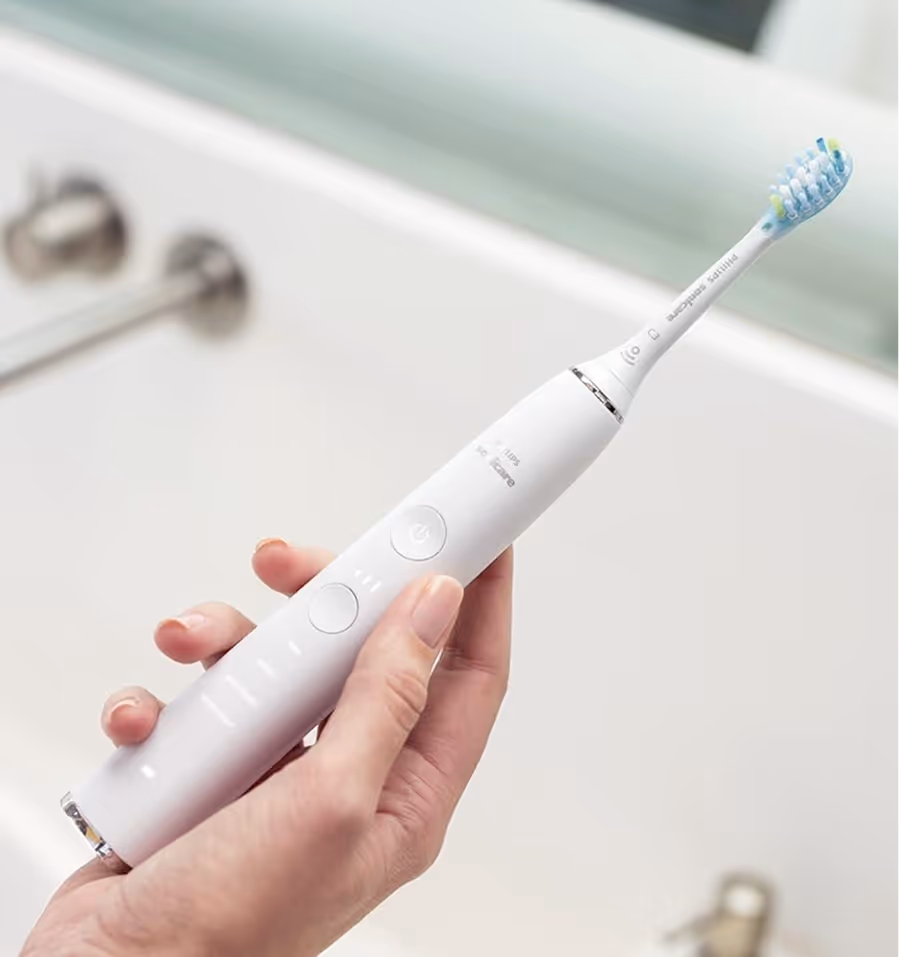 Hand holding a white electric toothbrush with blue and green bristles near a bathroom sink.