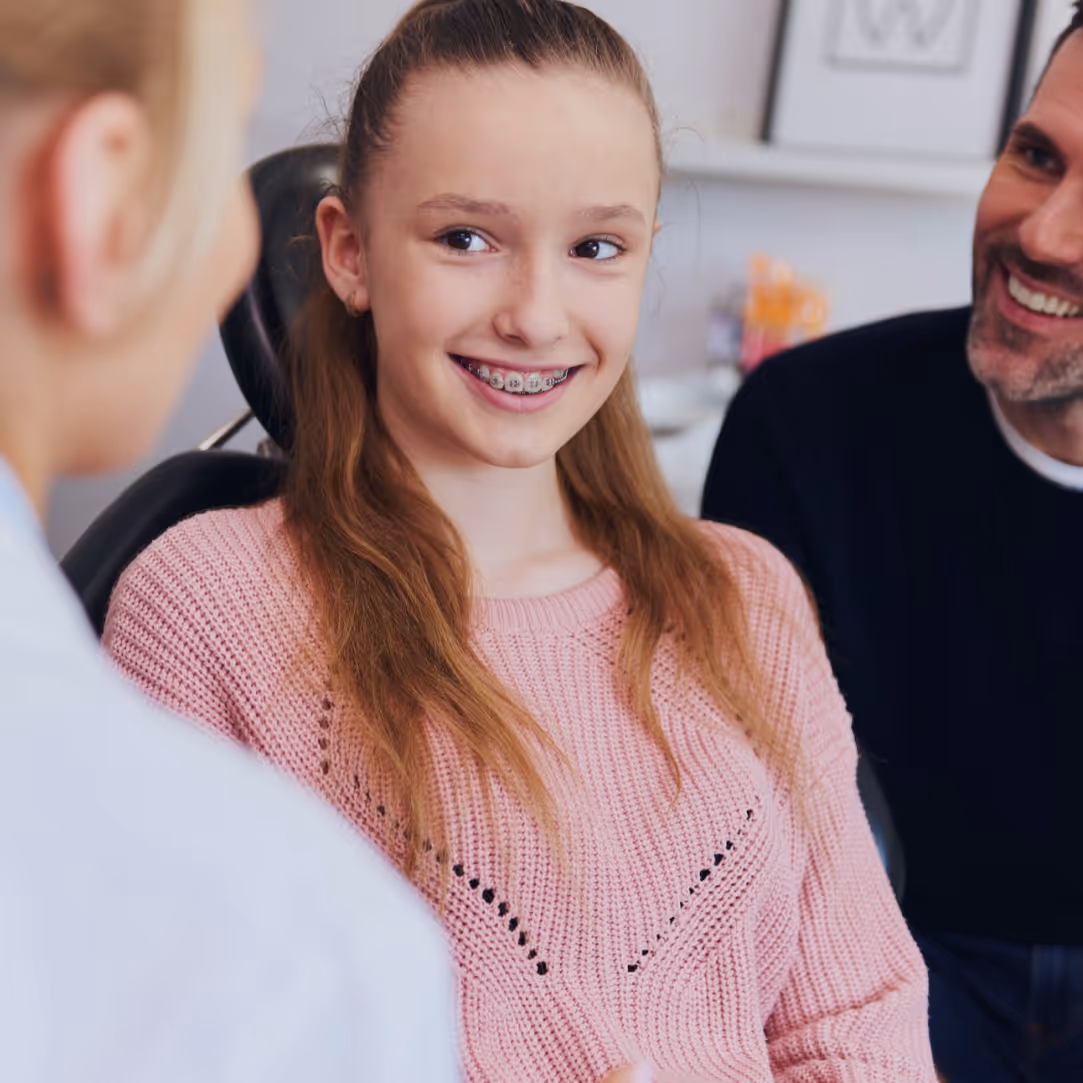 Smiling girl with braces in pink sweater sitting in a dental chair with an adult man and dentist.