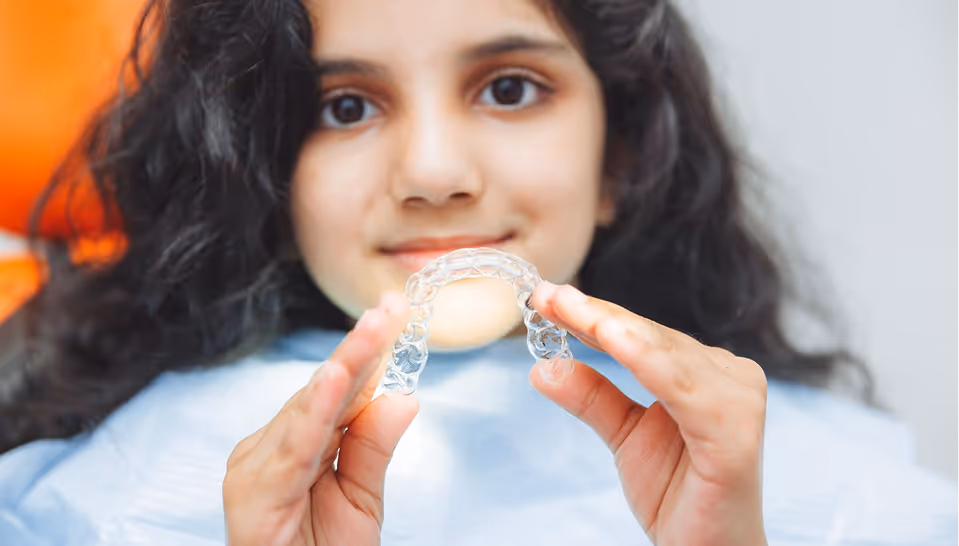 Young girl with dark curly hair holding a clear dental aligner with both hands.