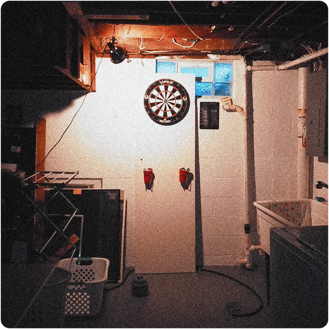 Dimly lit basement laundry area with dartboard on white door, laundry baskets, and utility sink.