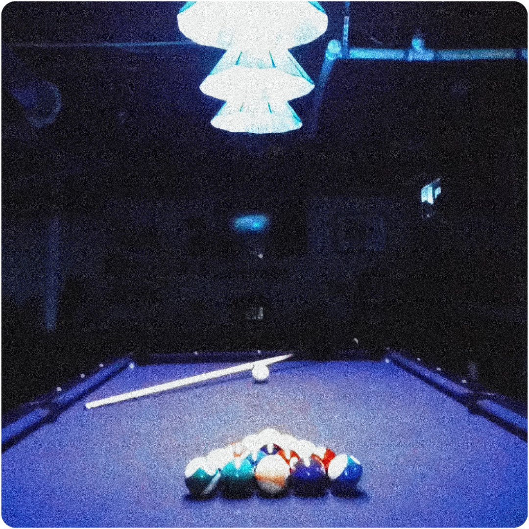 Pool table with billiard balls racked and cue stick resting on the table under a bright hanging light in a dark room.