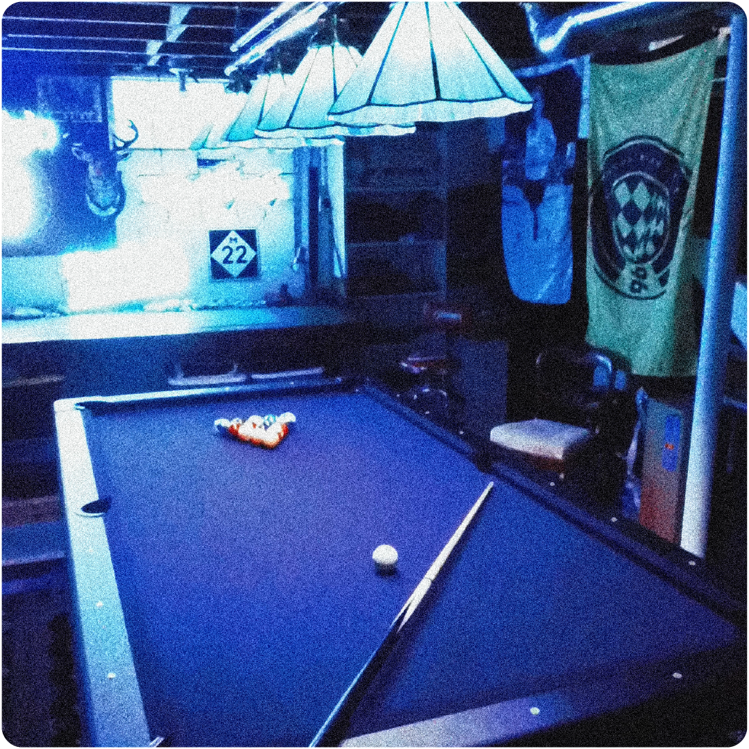 Dimly lit room with a pool table ready for a game, hanging lamps, a mounted deer head, and banners on the walls.
