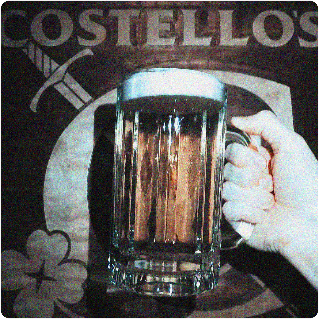Hand holding a glass mug of beer in front of a sign that reads 'Costello's' with a sword and clover graphic.