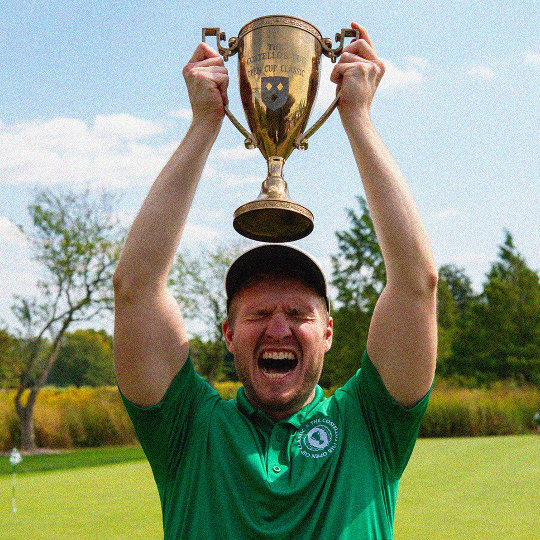 Man in green polo and black cap raising a gold trophy above his head with eyes closed and mouth open, celebrating outside on a golf course.
