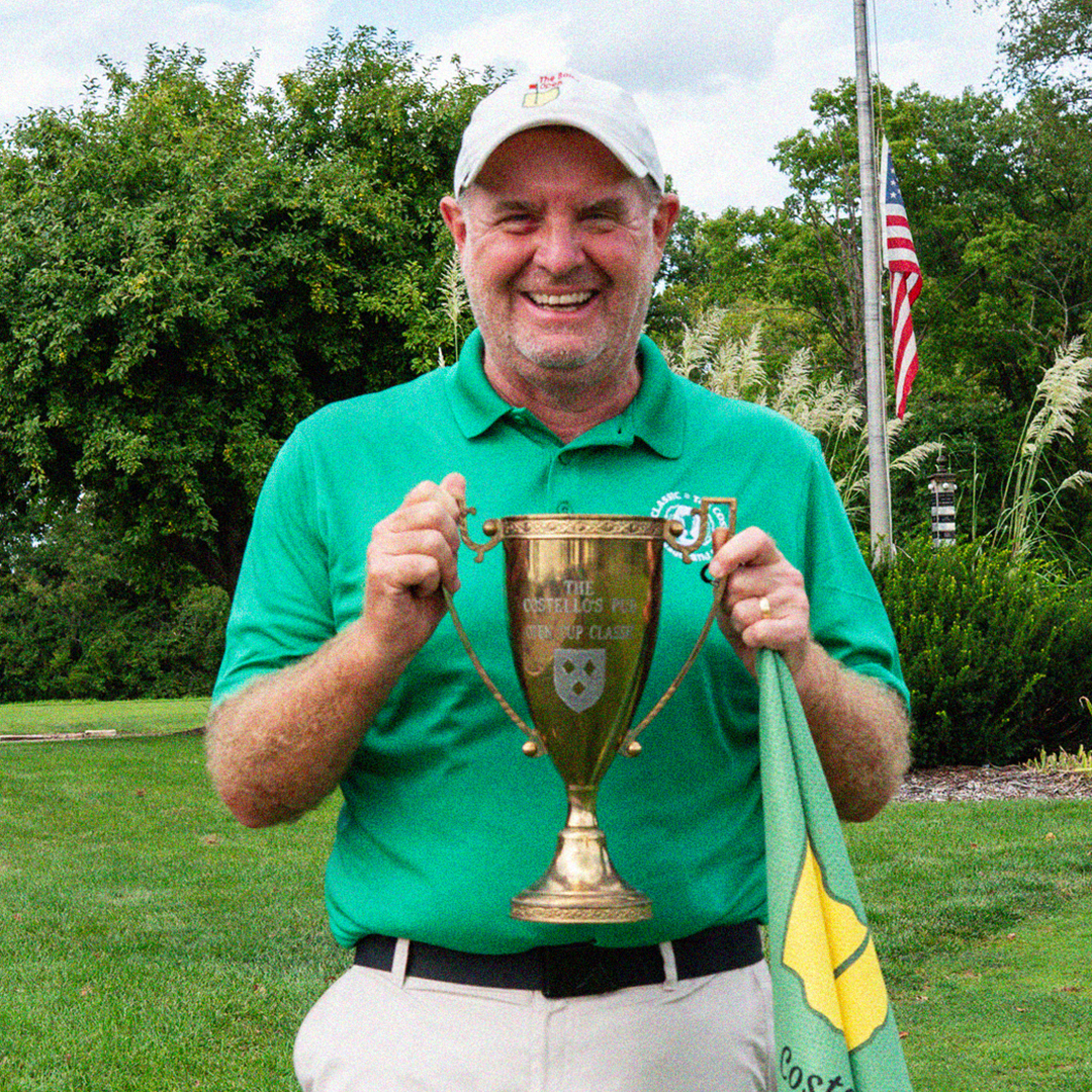 Smiling man in a green polo and white cap holding a trophy cup and a green flag outdoors with trees and an American flag in the background.