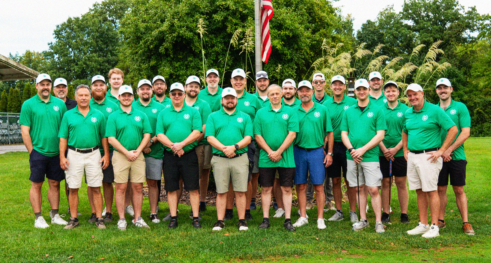Group of 25 men outdoors on green grass, all wearing matching green polo shirts and white caps, standing in front of tall plants with an American flag on a pole behind them.