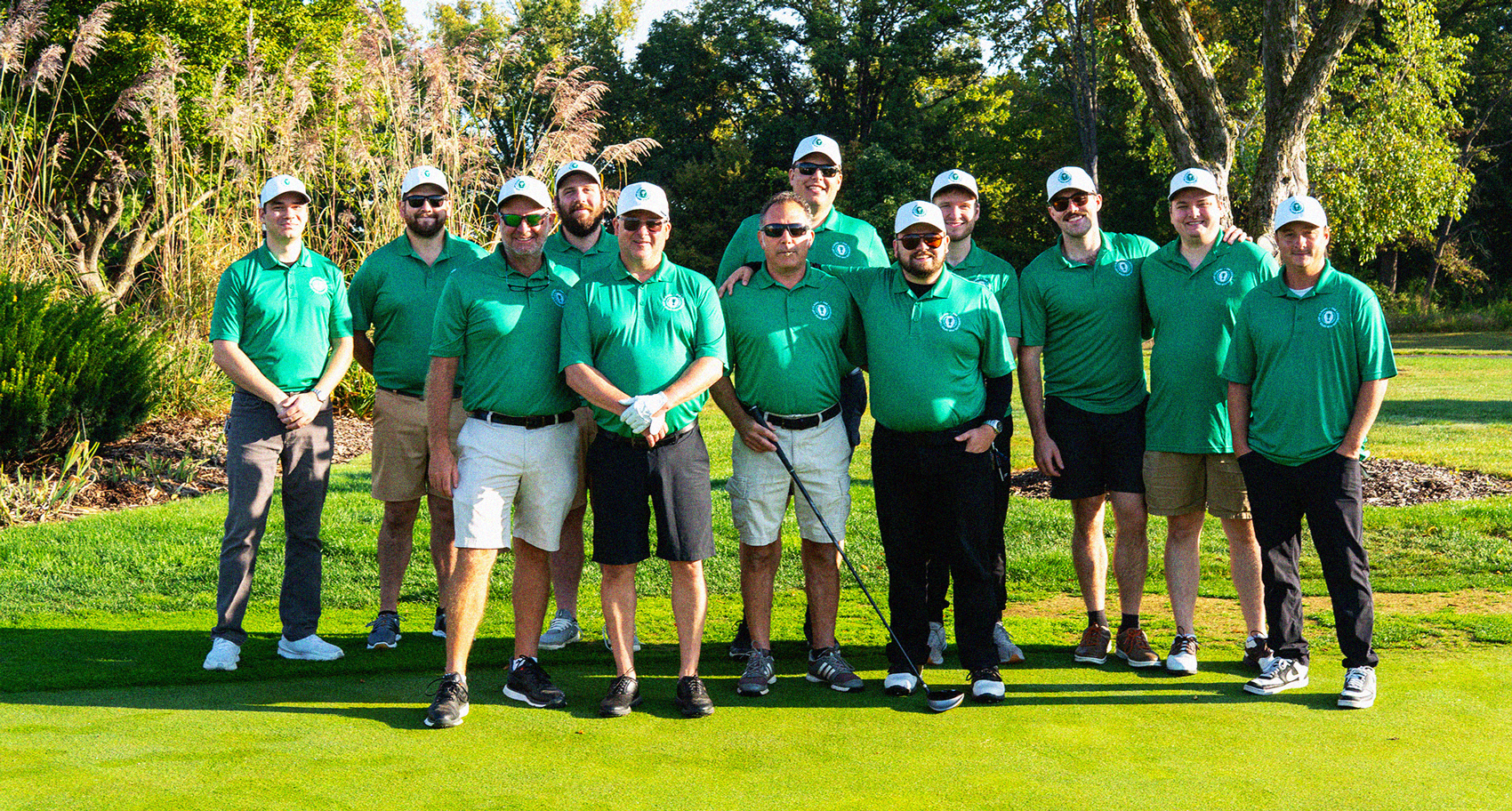 Group of eleven men standing on a golf course green wearing matching green polo shirts and white caps, with trees and tall grass in the background.