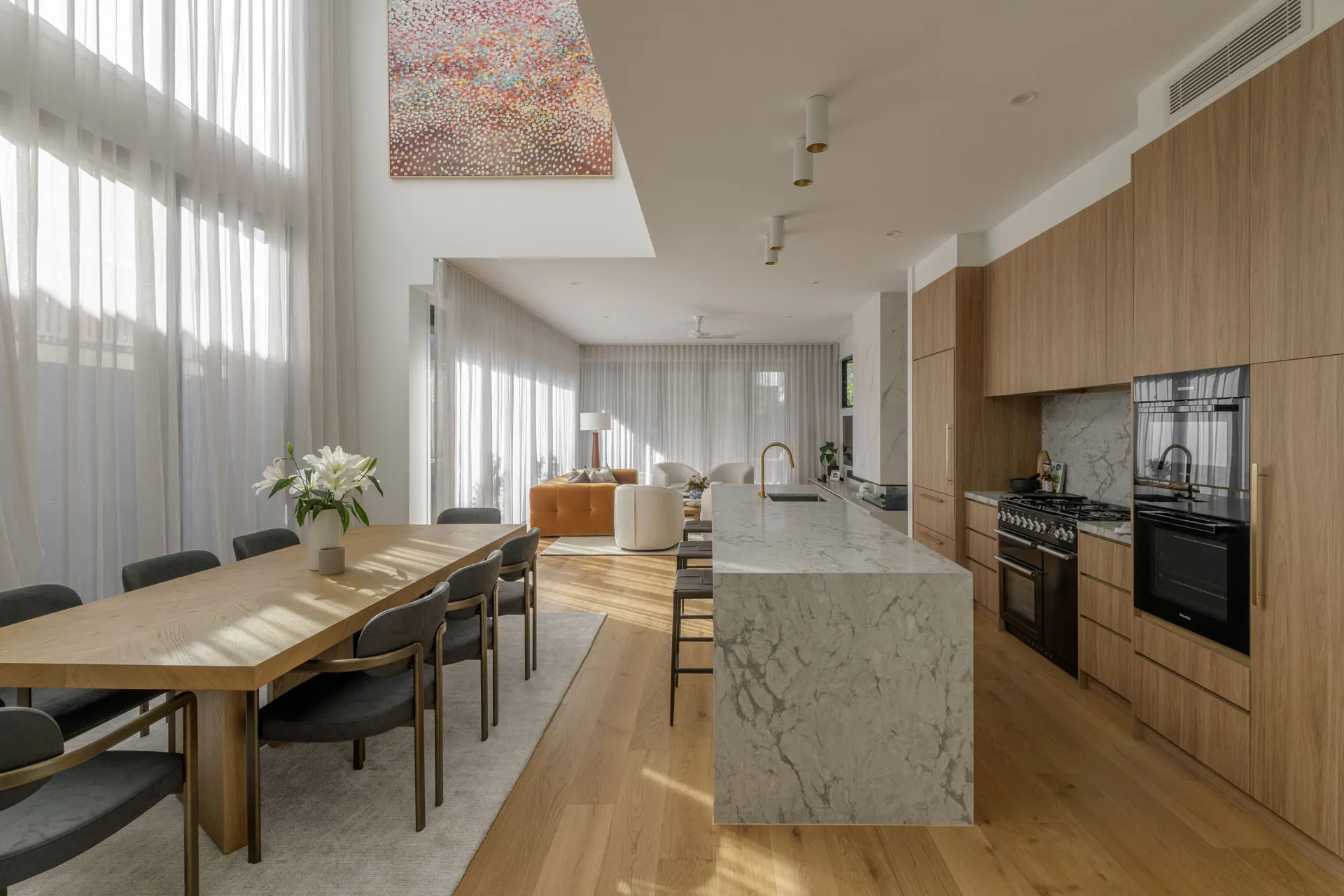 Modern open-plan kitchen and dining area with marble island, wooden cabinets, and large windows with sheer curtains.