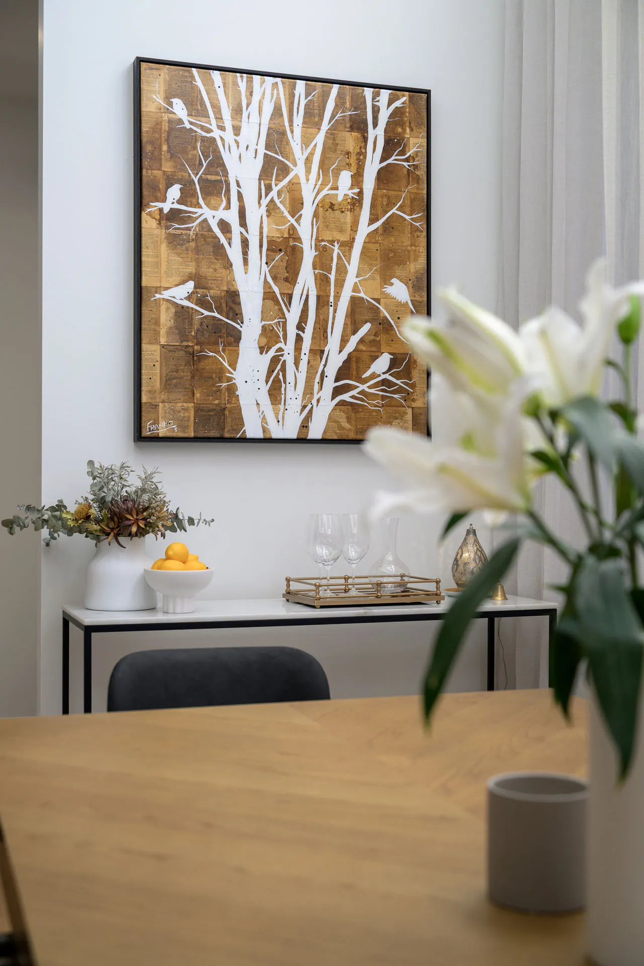 White tree and birds artwork on wall above console with vase, bowl of lemons, and glassware tray.
