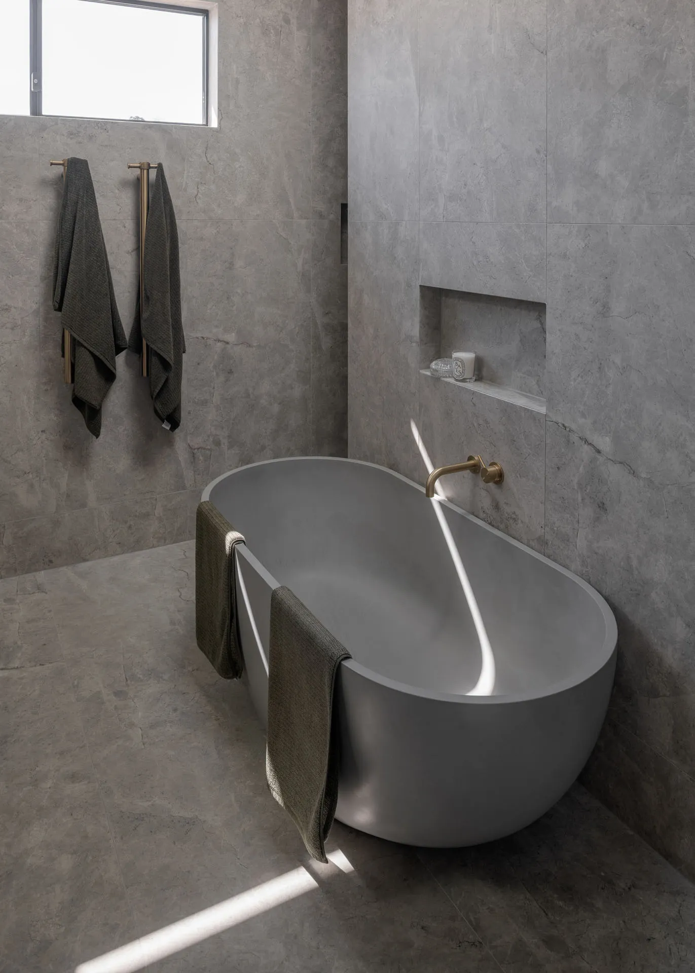 Modern gray bathroom with freestanding oval tub, bronze faucet, towels, and stone tile walls and floor.