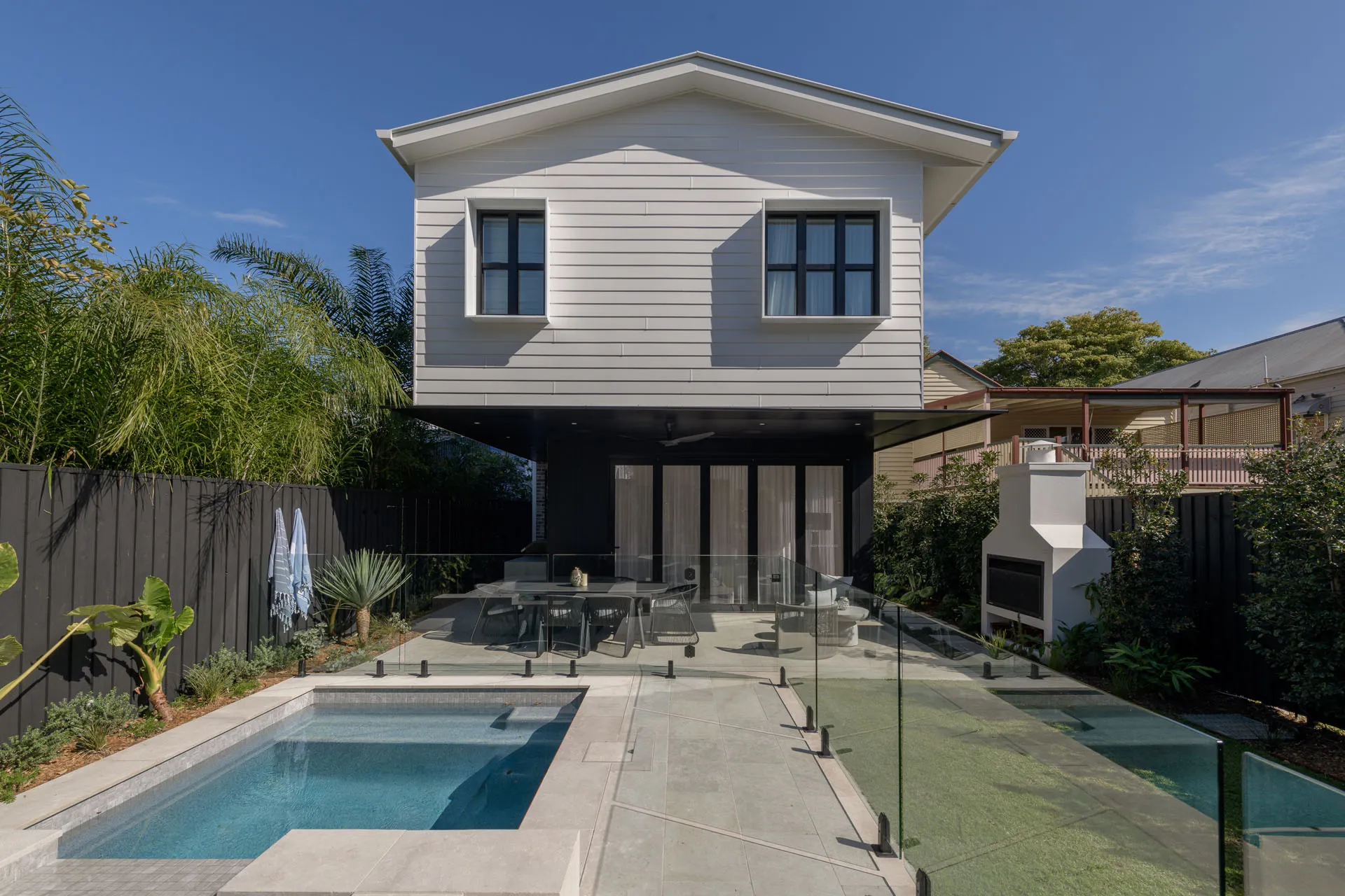 Modern two-story house with white siding, glass fence, outdoor pool, patio furniture, and built-in grill.