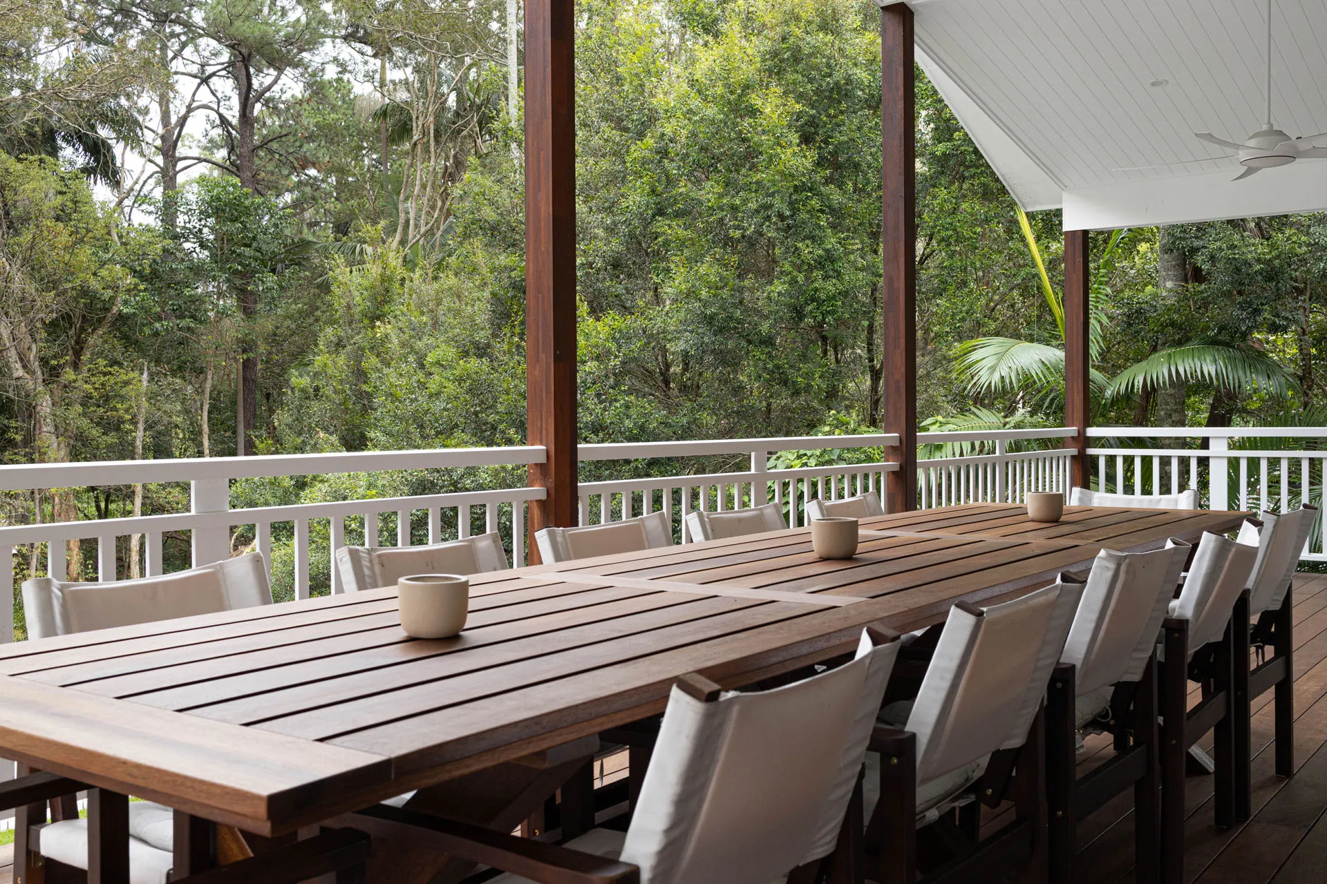 Long wooden outdoor dining table with white cushioned chairs on a covered patio surrounded by greenery.