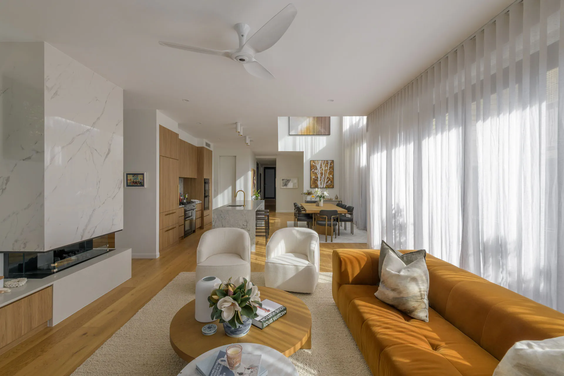 Bright modern living room with orange sofa, white chairs, wood floor, kitchen, and large sheer curtains.