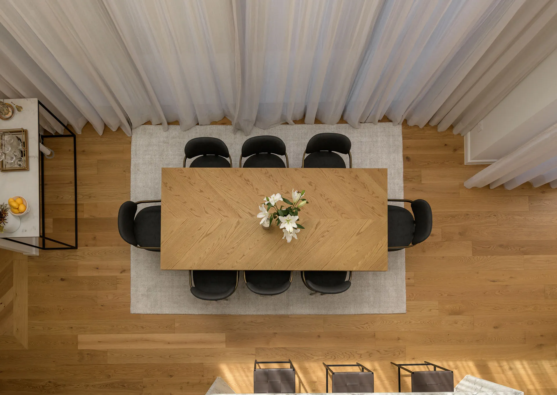 Top view of a wooden dining table with six black chairs and a vase of white flowers on a light rug.