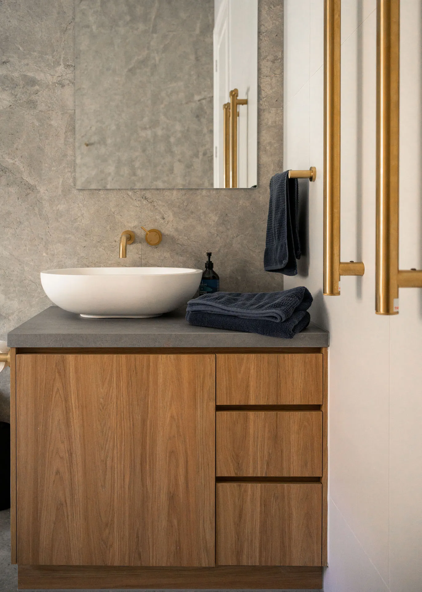Modern bathroom vanity with wooden cabinet, white vessel sink, gold faucet, and navy towels.