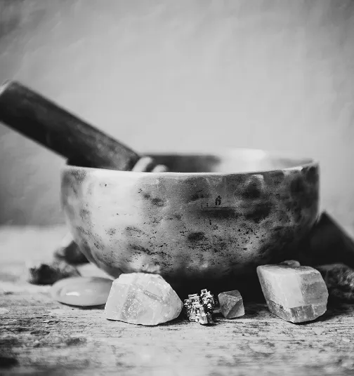 Black and white photo of a rustic metal singing bowl with a wooden striker, surrounded by various crystals on a textured wooden surface.