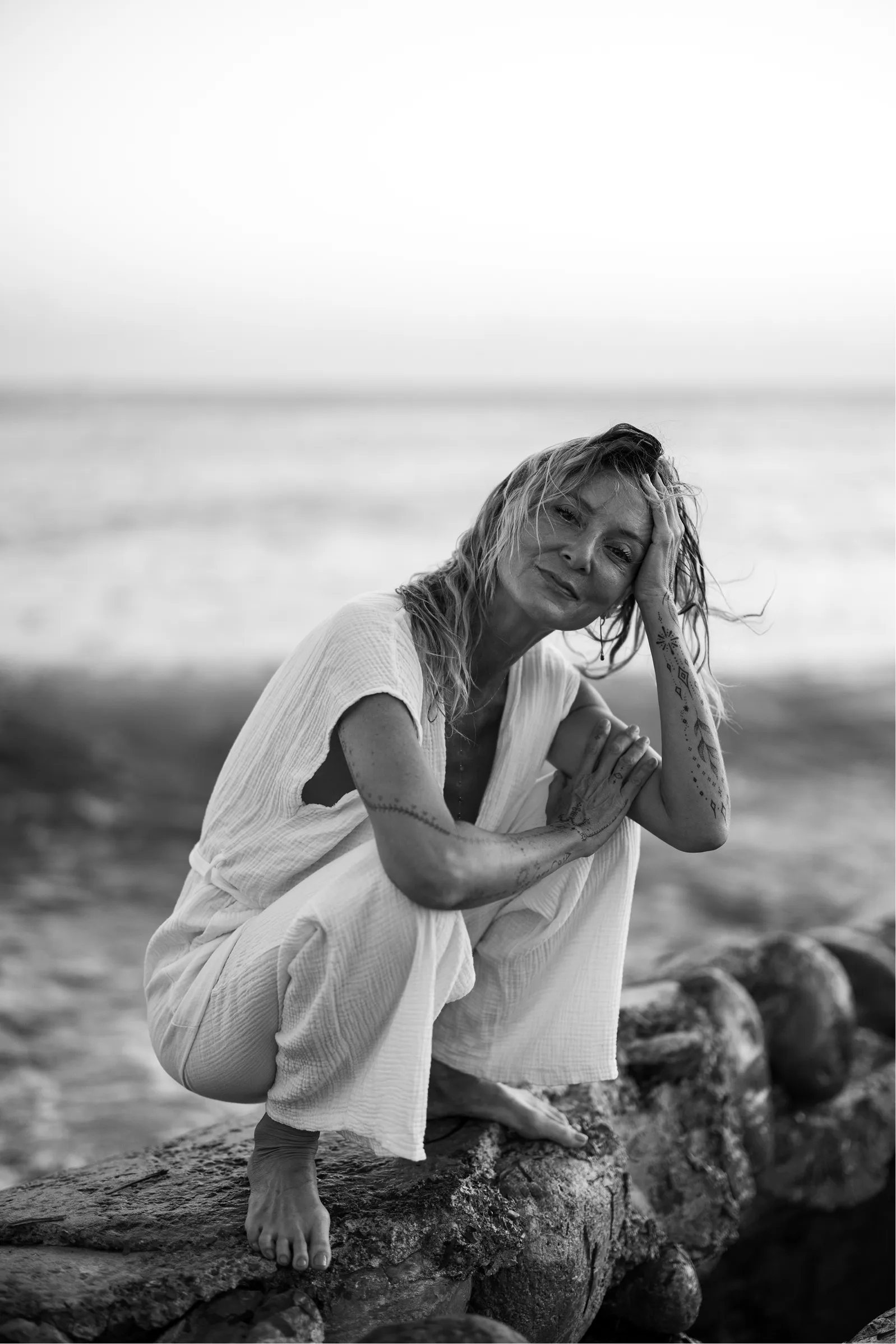 Black and white photo of a woman in white clothing crouching barefoot on rocks near the ocean, with windblown hair and tattoos on her arms.