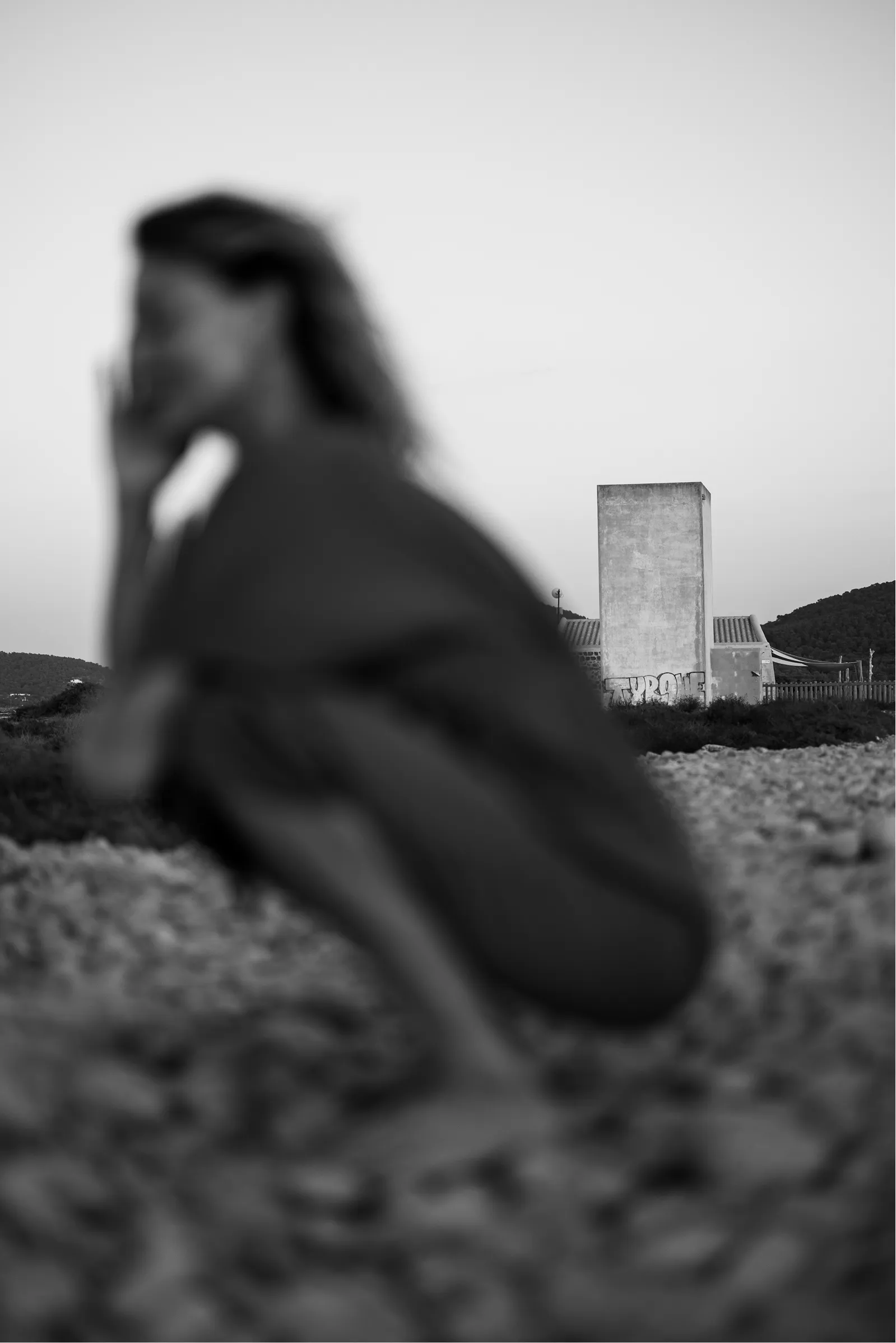 Black and white image of a person squatting on rocky ground with a tall, rectangular concrete structure in the background.