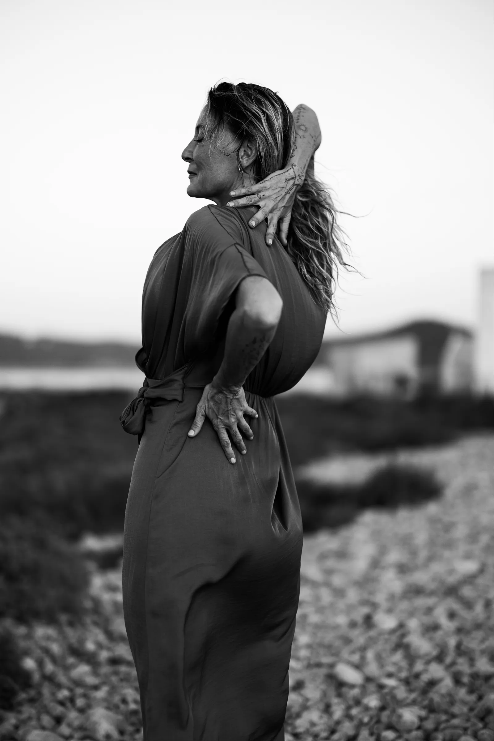 Black and white photo of a woman standing on a rocky path with eyes closed, one hand touching her neck and the other on her lower back.