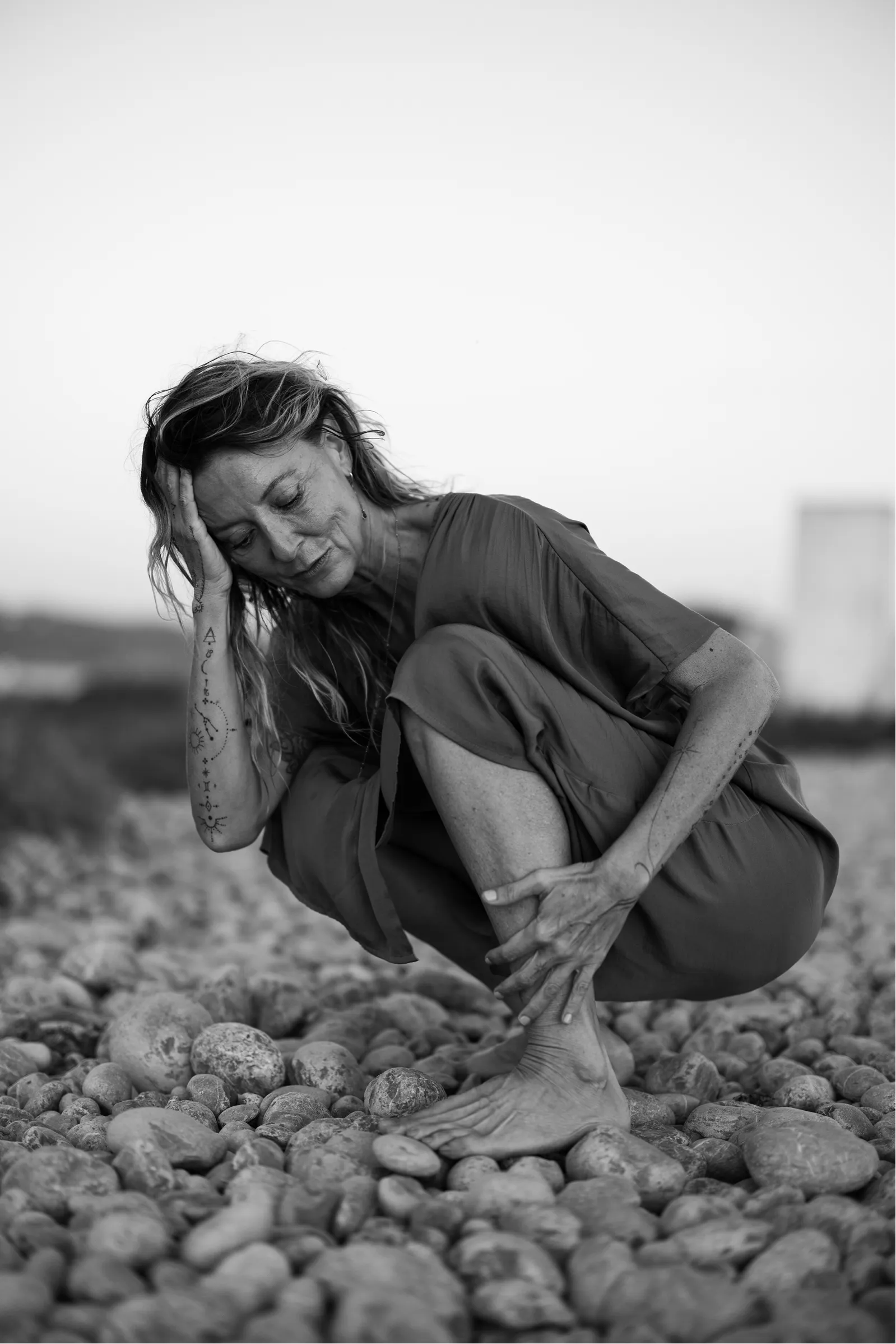 Black and white photo of a barefoot woman with tattoos crouching on rocky ground, resting her head on her hand with eyes closed.