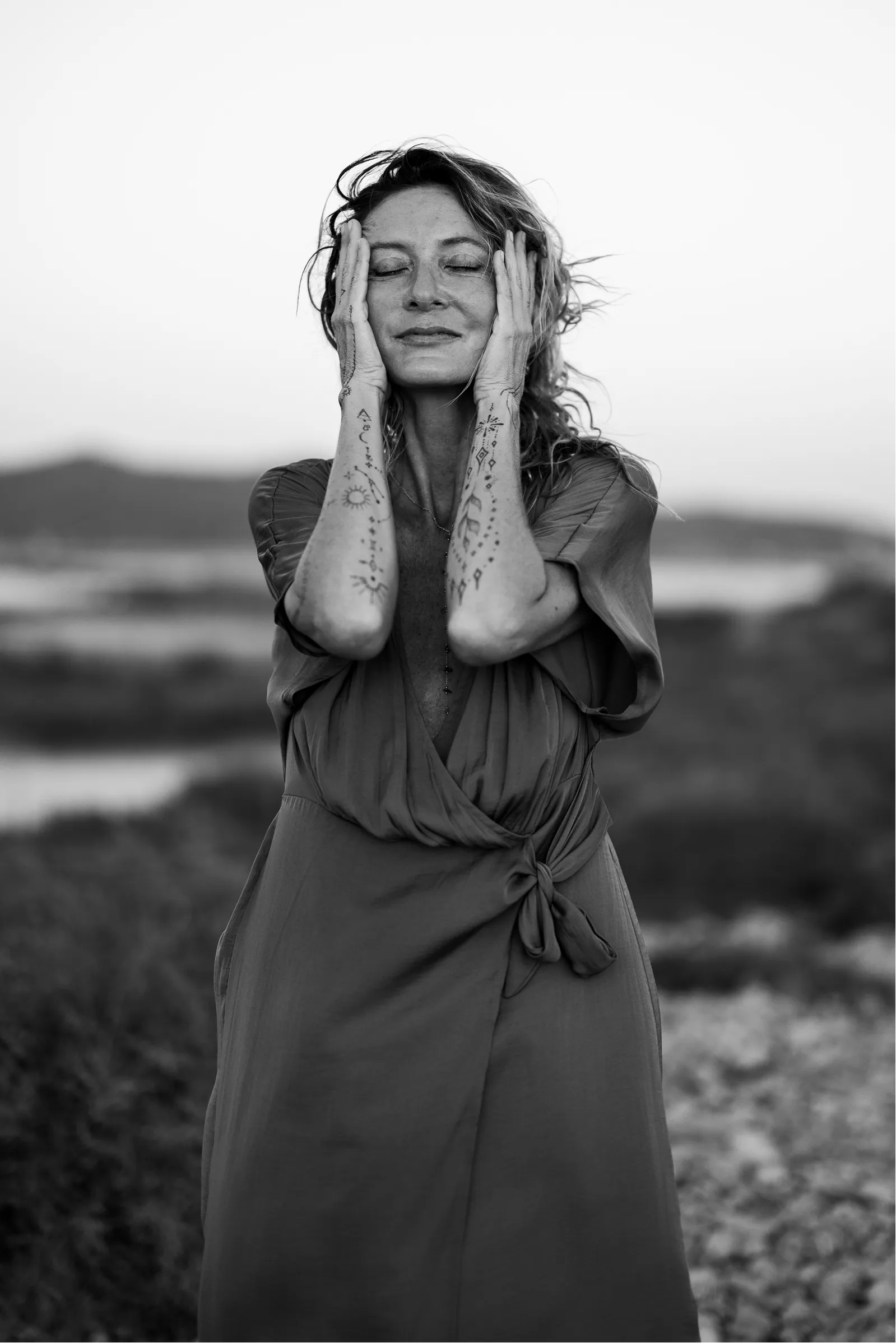 Black and white image of a woman with closed eyes touching her face with tattooed forearms, standing outdoors with a blurred natural background.