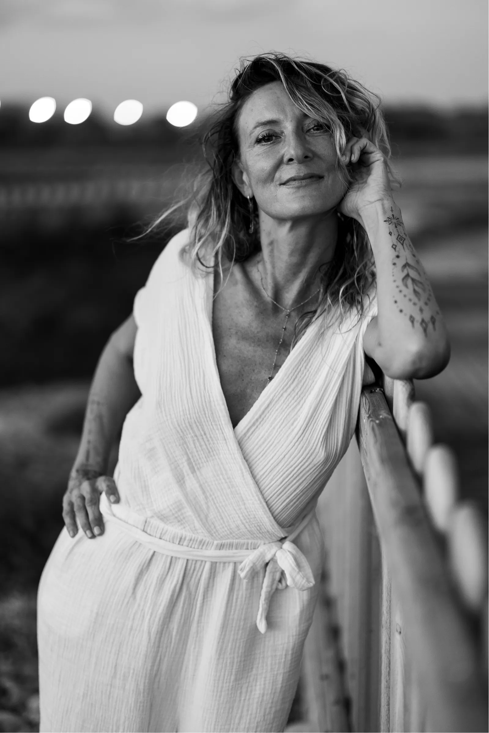 Black and white portrait of a woman leaning on a wooden railing, wearing a textured white dress with tattoos visible on her left arm.