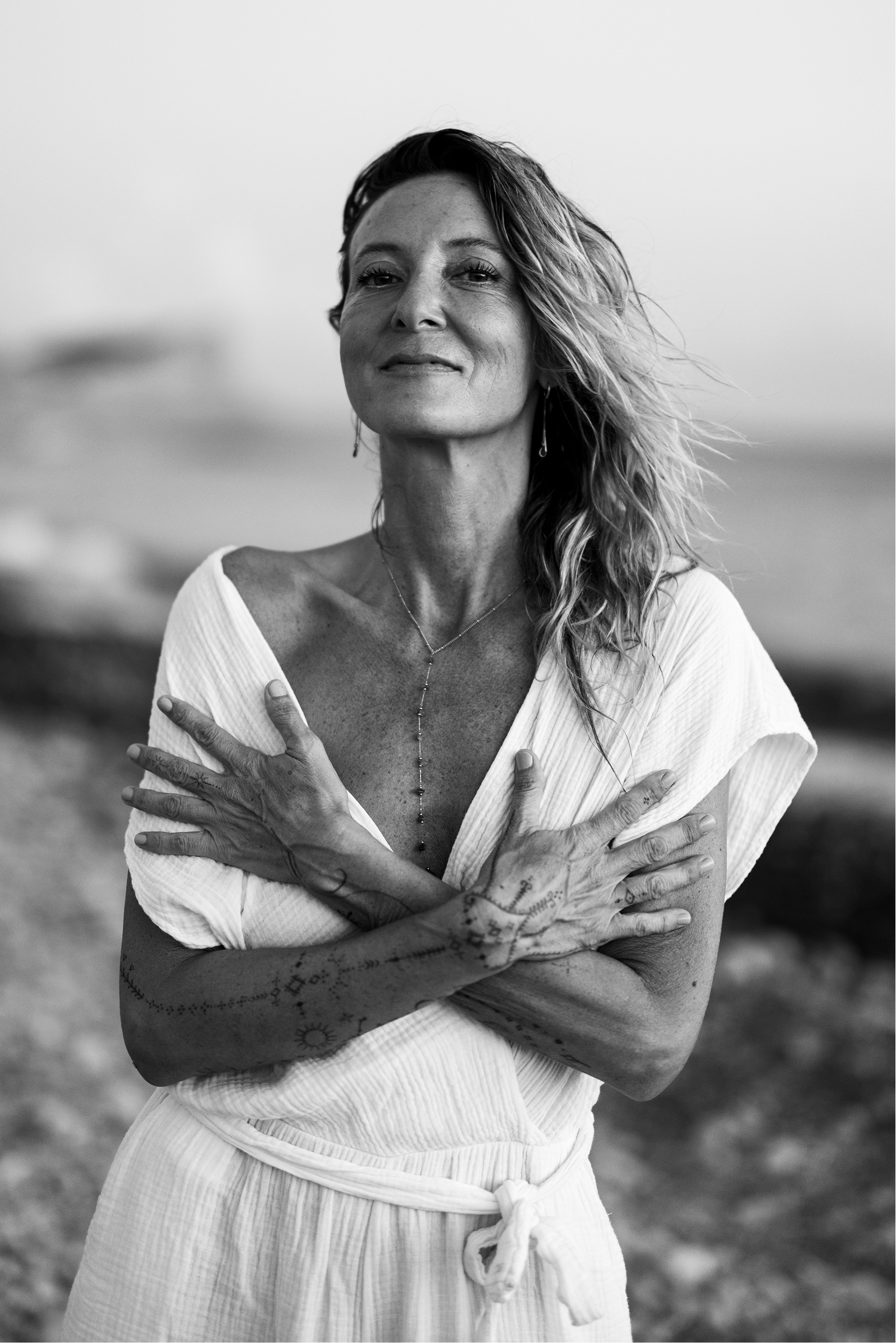 Black-and-white portrait of a woman with long wavy hair smiling slightly, wearing a light dress with crossed tattooed arms.