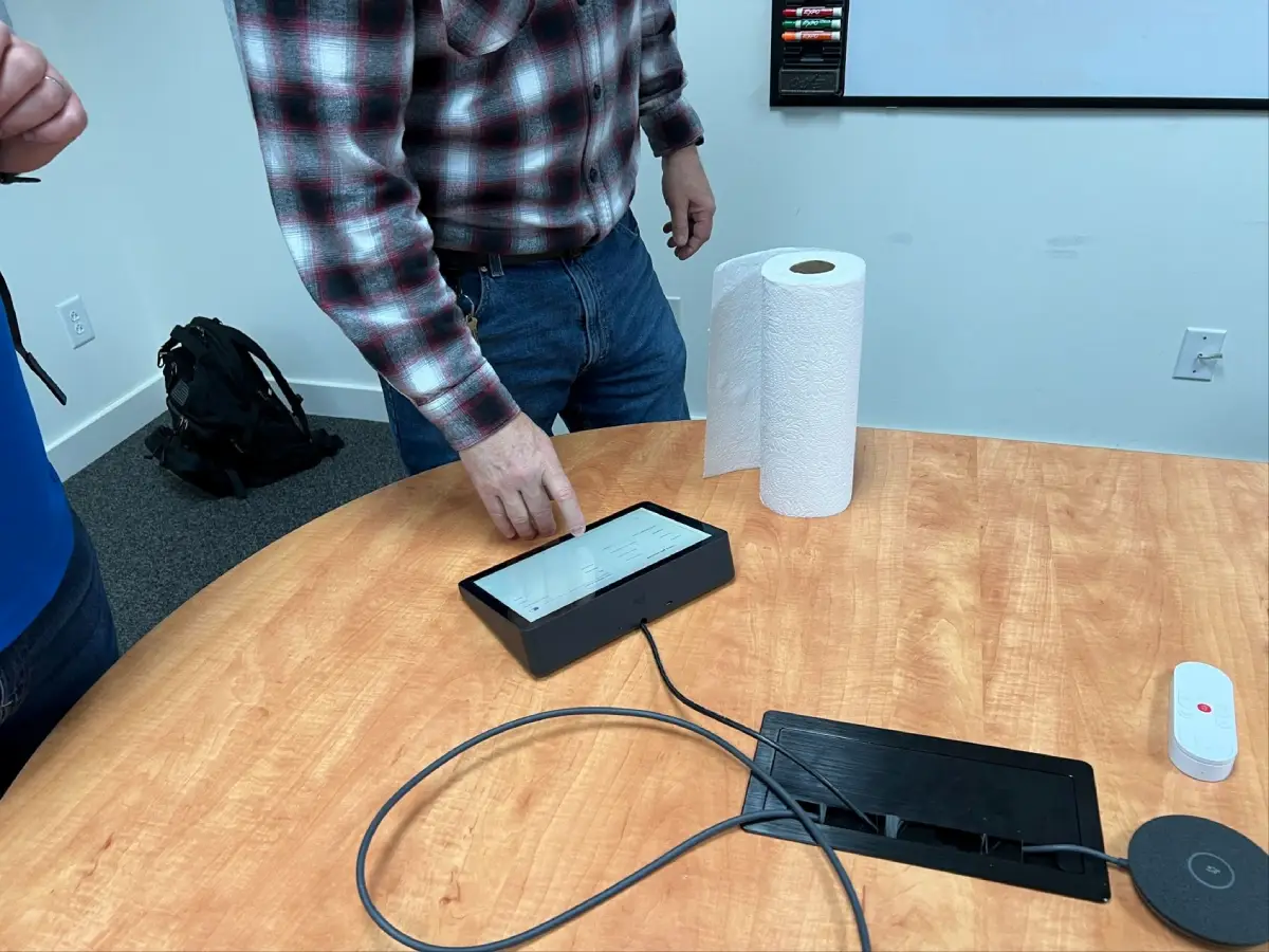 Hand using a conference room touch controller on a round table with a table microphone and cable cubby