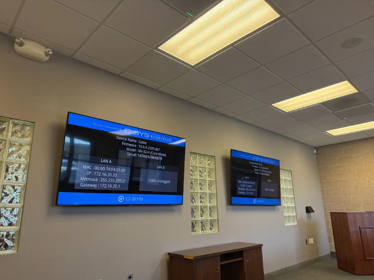 Two wall displays in a meeting room showing the Q-SYS system status screen