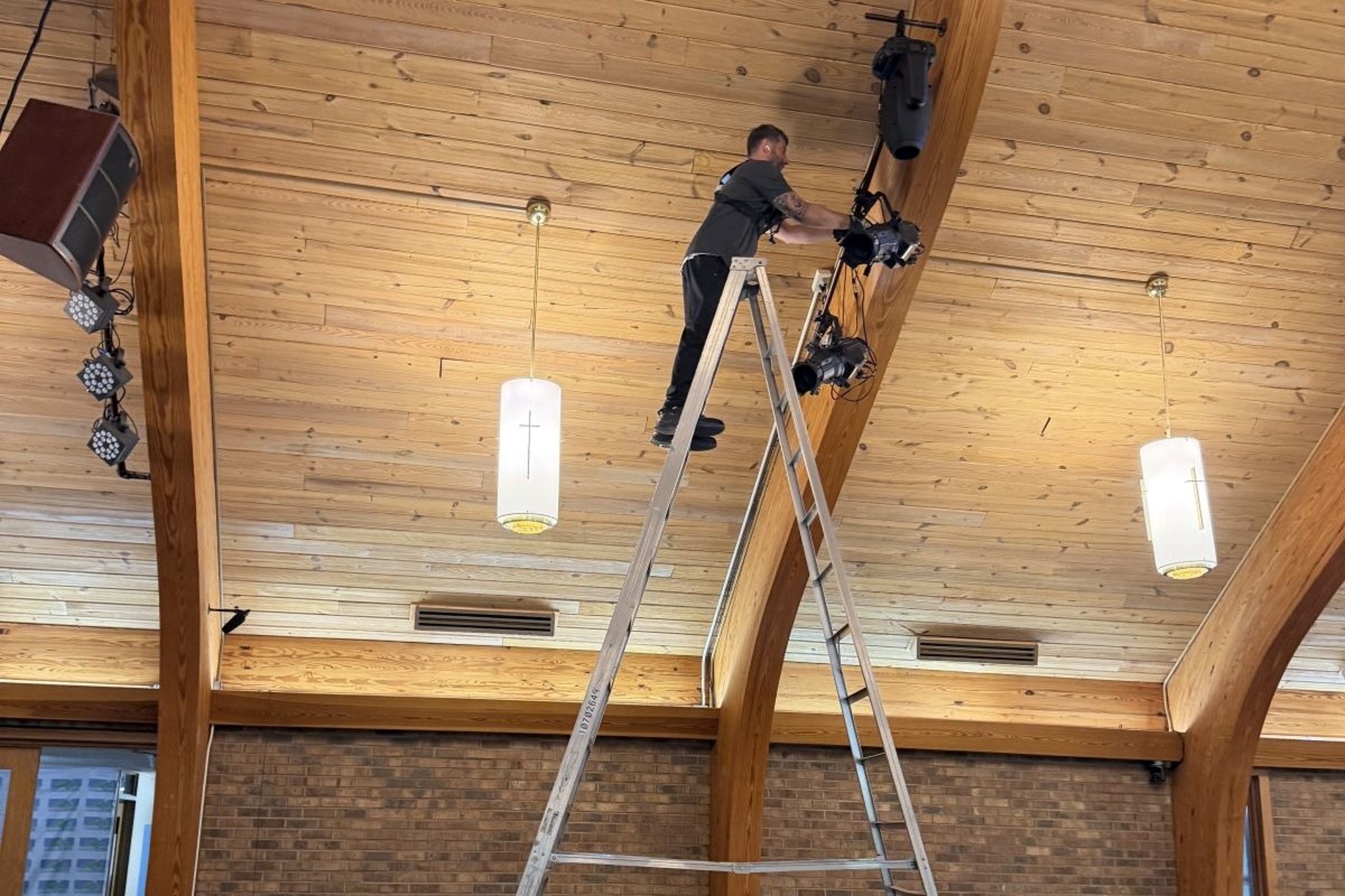 RYGID AV technician on a ladder working on lighting fixtures mounted to a wooden beam in a church sanctuary