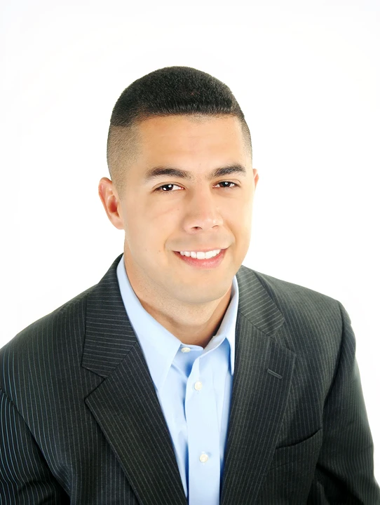 Smiling man with short dark hair wearing a black pinstripe suit jacket and light blue collared shirt on a white background.