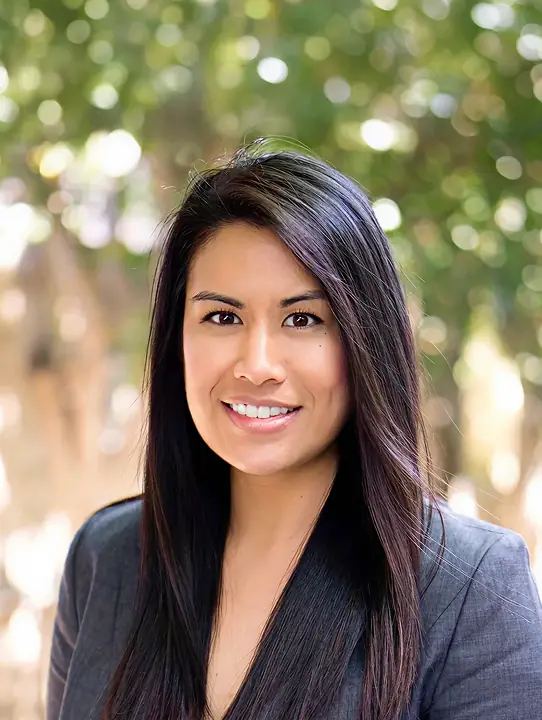 Smiling woman with long dark hair wearing a gray blazer, standing outdoors with blurred green foliage background.