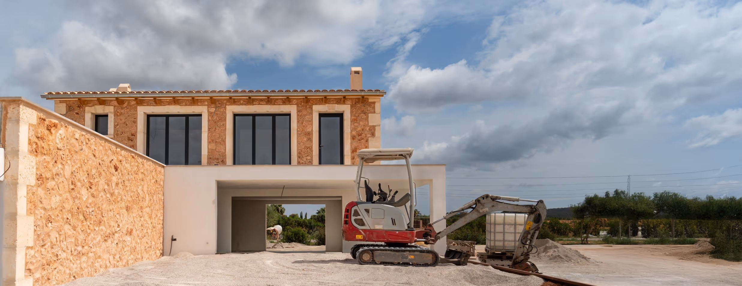 Construction or renovation site of a modern house featuring a stone facade on the second floor and a white, open ground level with a large archway. A small red and white excavator is parked on the gravel in front of the structure.