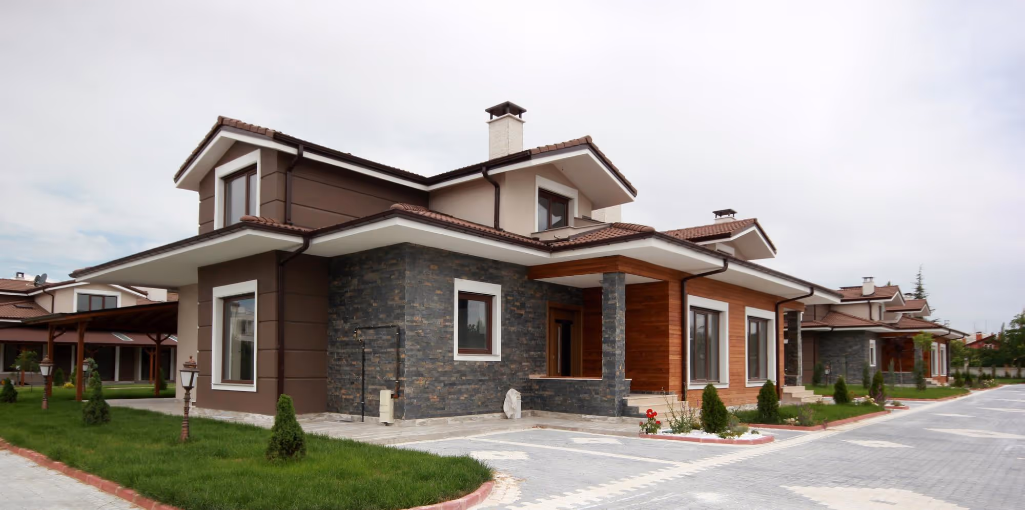 Exterior view of a modern, multi-textured suburban house in a residential row, featuring a brown and beige painted facade, dark gray stacked stone cladding, and some light wood paneling, with a paved driveway and small landscaped front yard.