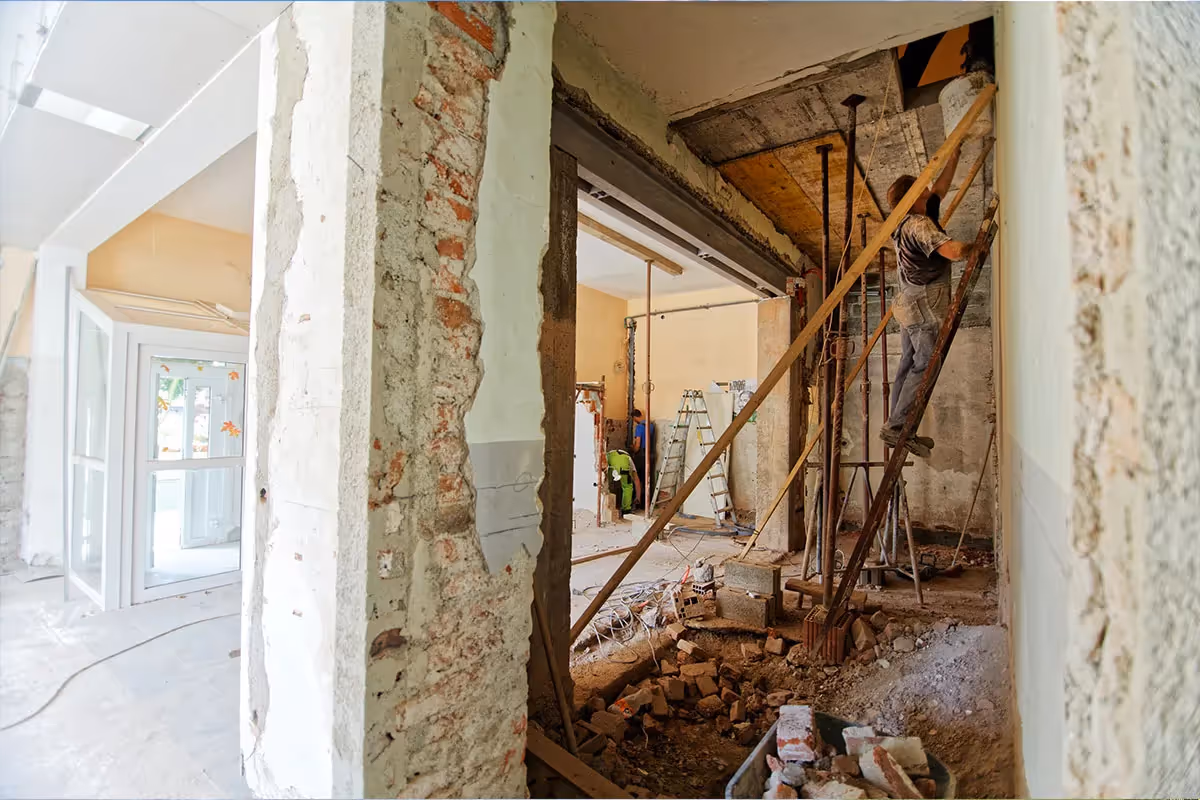 Interior view of an apartment or building undergoing major renovation or demolition, showing exposed brick walls, construction debris, and a worker using temporary wooden shoring to support a ceiling beam. Other workers are visible in the background.
