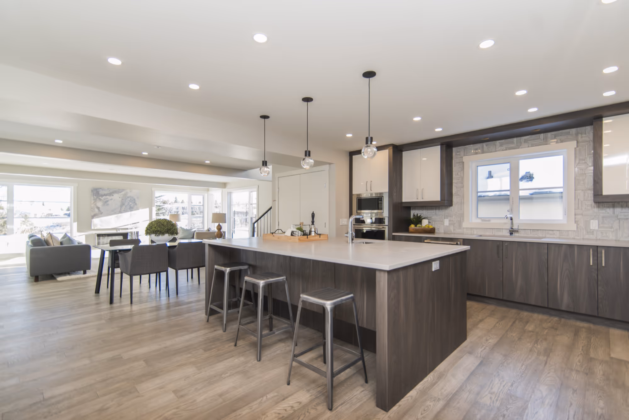 Modern open-concept kitchen and dining area featuring a large island with white quartz countertop and three gray metal stools, dark wood grain lower cabinets and island, and white upper cabinets. The adjacent dining area has a dark table and chairs.