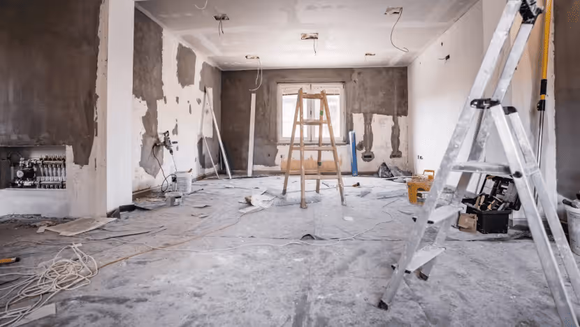 Interior of a room undergoing renovation, with walls partially plastered or patched, exposed wiring, and a concrete floor covered with dust sheets. A wooden stepladder is positioned in the center, and a metal ladder is visible in the foreground on the right.
