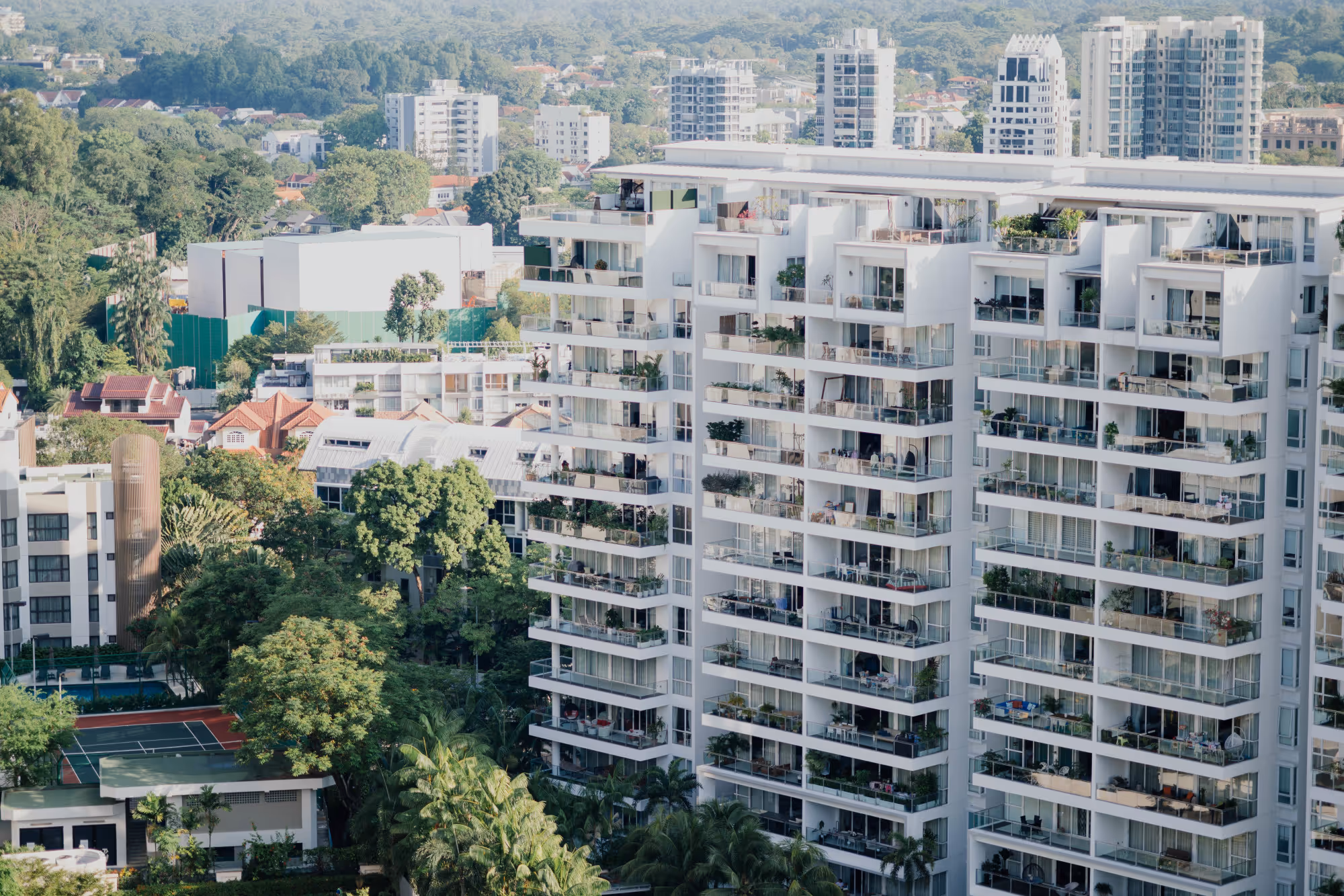 High-angle view of a dense urban residential area, dominated by a large, modern white apartment building with numerous balconies and greenery, surrounded by lower-rise buildings and abundant tropical trees.