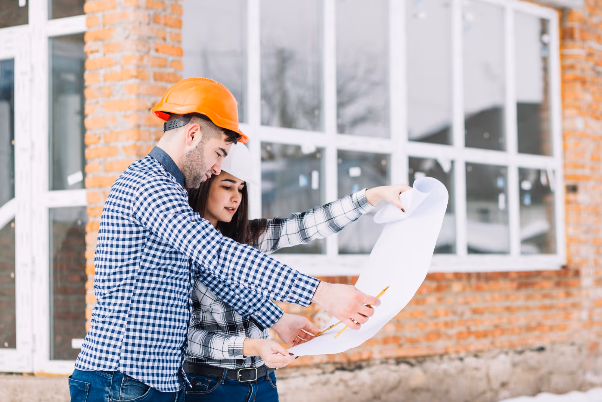Two people, a man and a woman wearing construction hard hats, are standing outside a building with a brick facade and large windows, looking down and discussing architectural blueprints they are holding.