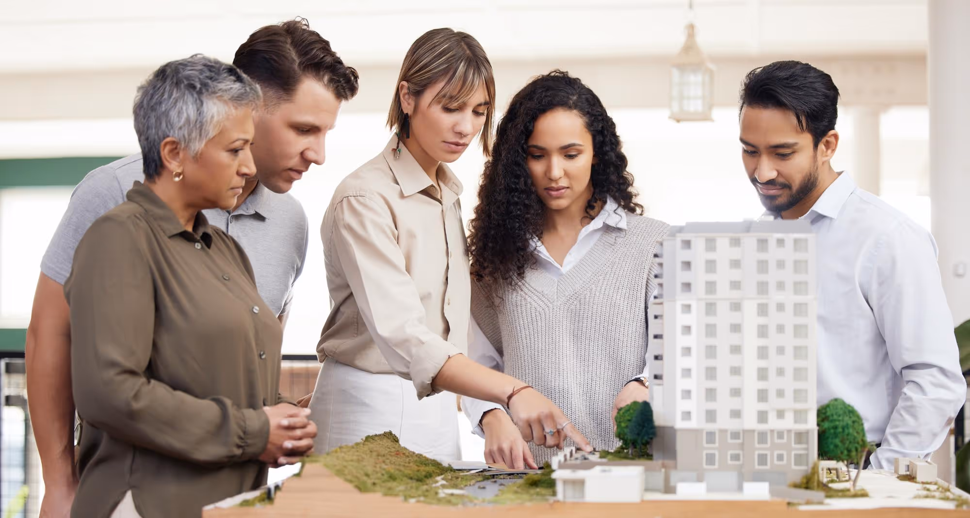 Five diverse architects or project managers are gathered around a detailed architectural model of a multi-story building and surrounding landscape, pointing and discussing the design.