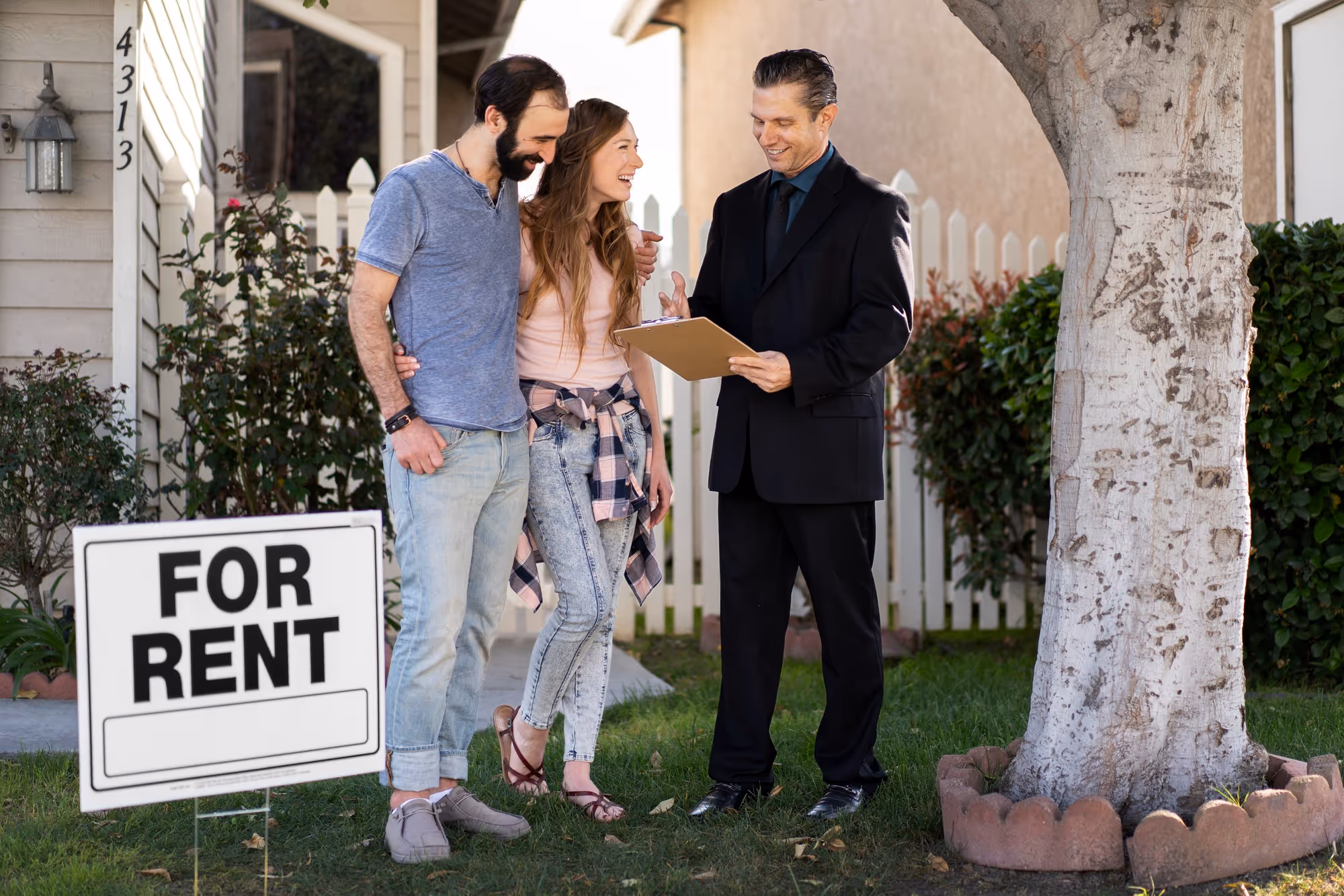A real estate agent in a black suit showing a clipboard to a smiling young couple standing in the front yard of a suburban house, next to a large tree and a 'FOR RENT' sign.