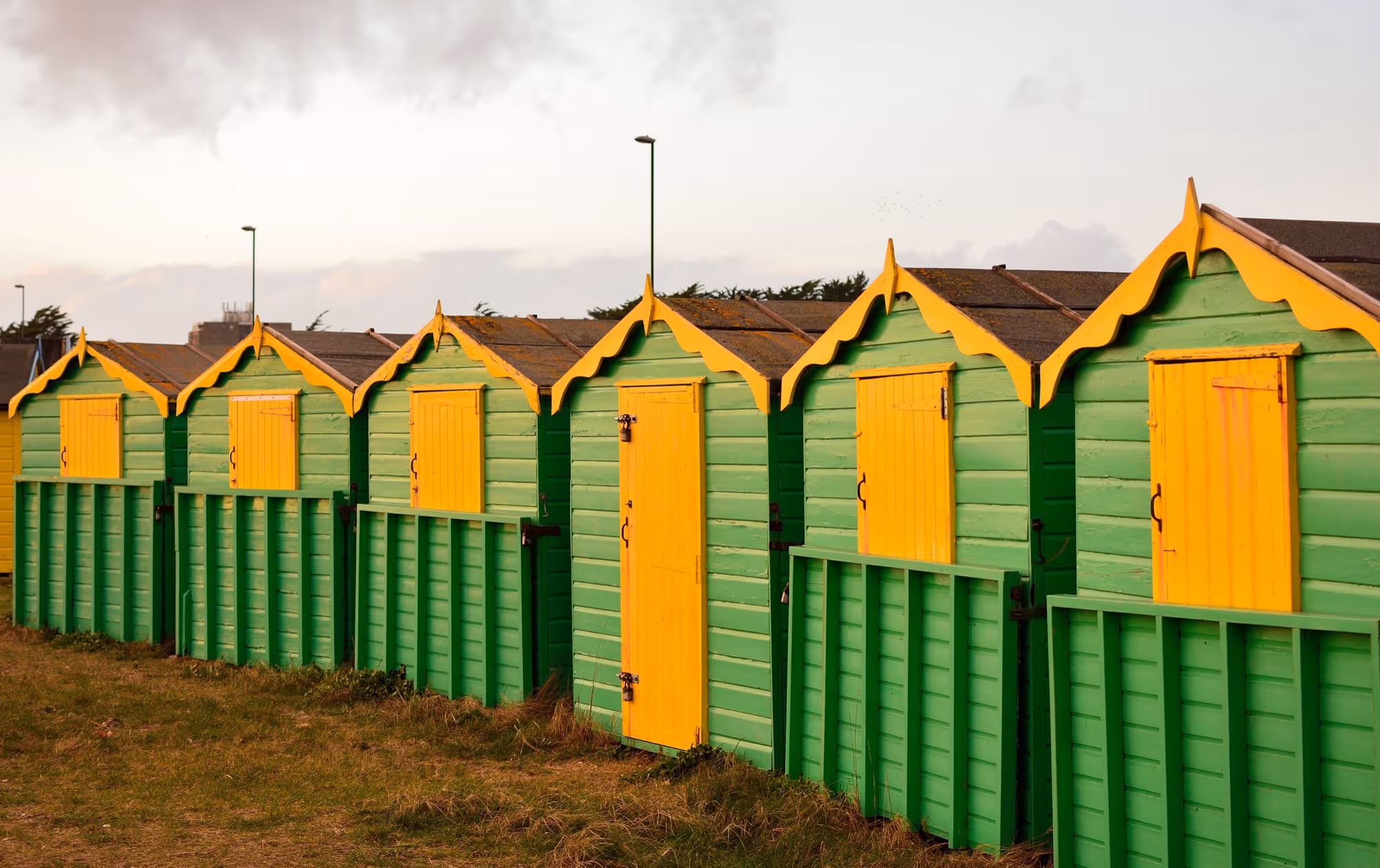 A row of traditional beach huts painted in vibrant lime green with contrasting bright yellow trim, doors, and window shutters, under a pale, overcast sky.