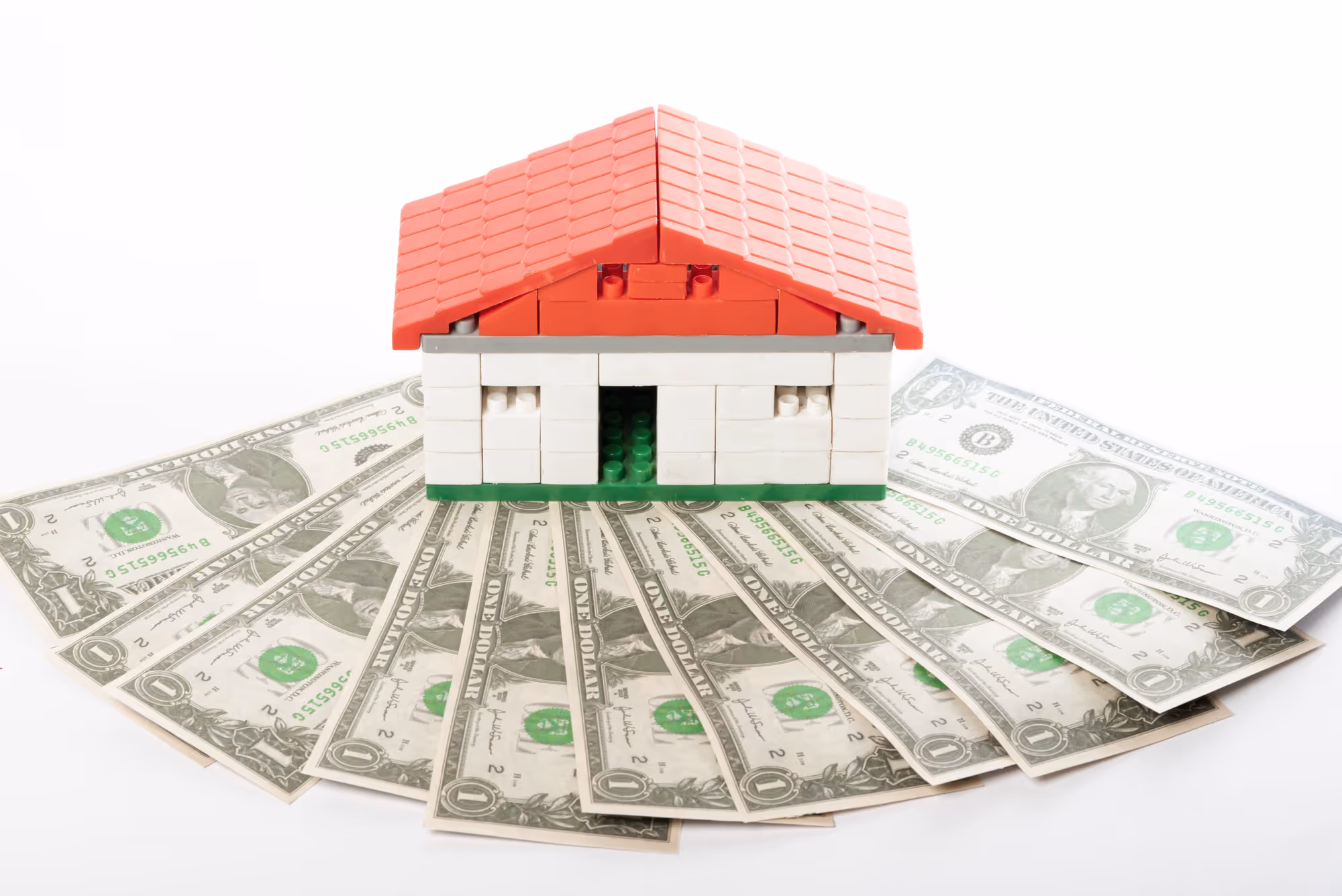 A small house constructed from red, white, and green plastic toy building blocks (like LEGO), sitting on a semi-circle fan of one-dollar bills, against a white background, symbolizing the cost of a home.