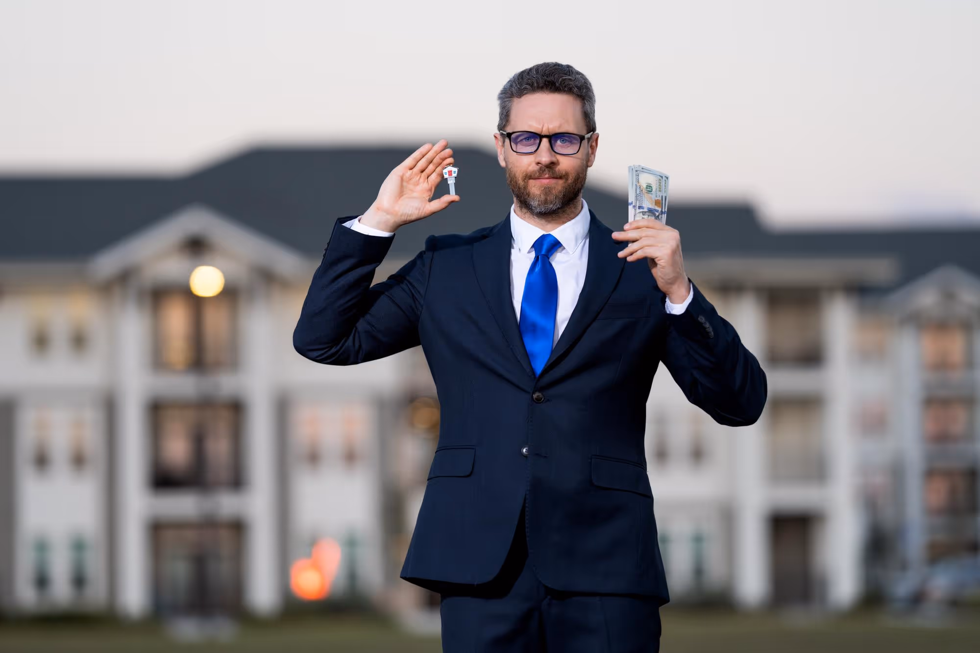 Serious real estate agent or businessman in a dark suit and blue tie holding a house key in one hand and a stack of money in the other, standing outside with a large apartment or townhouse complex blurred in the background.