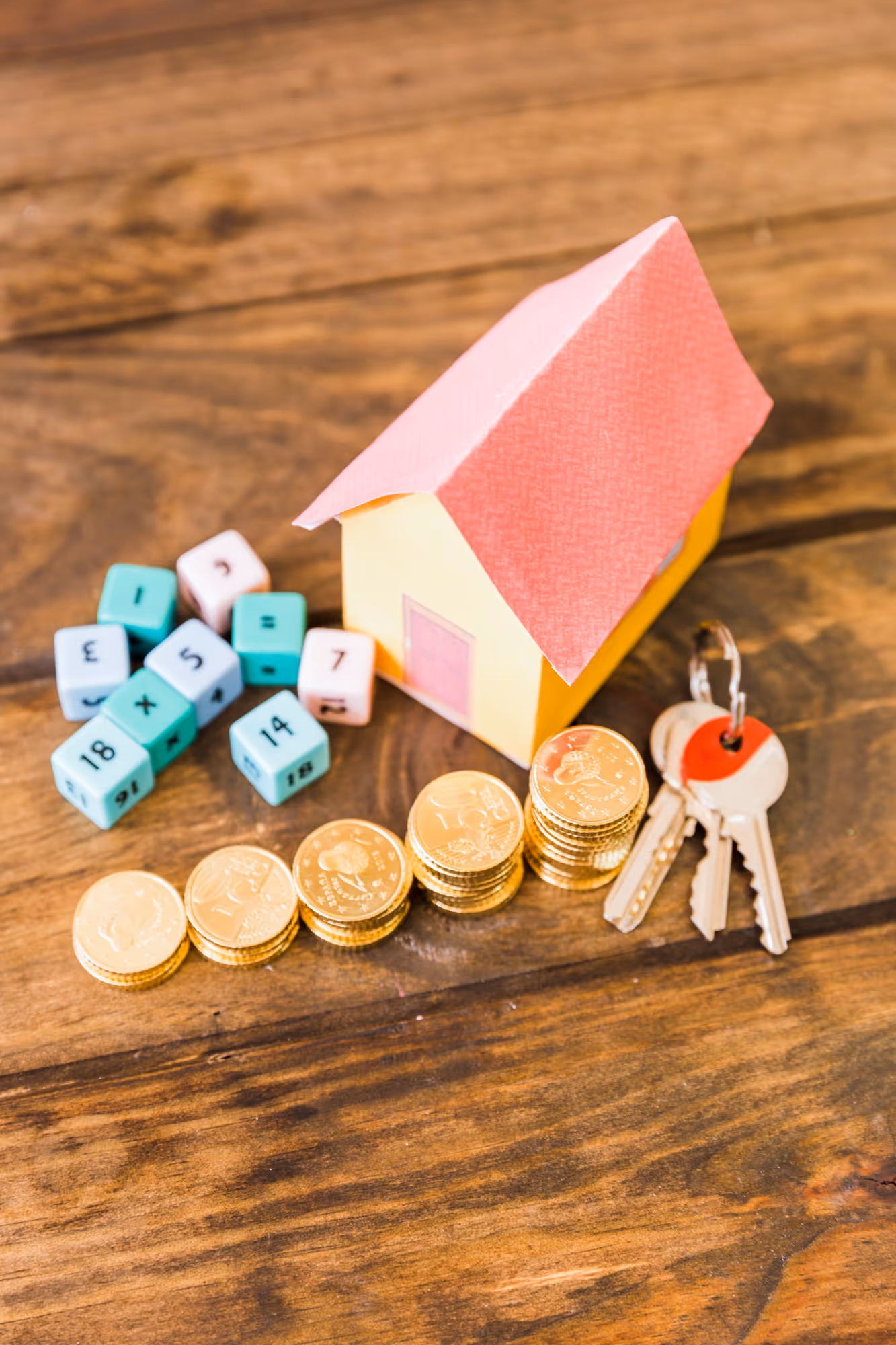 Still life of real estate investment concept with a small paper house model, stacks of gold coins, a set of keys, and scattered small math dice on a rustic wooden surface.
